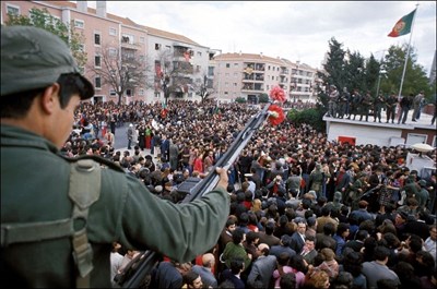 A person holds a gun with a carnation flower in the barrel overlooking a crowd.