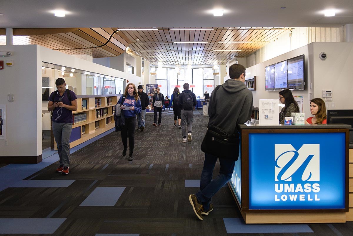 Interior of O'Leary Library with UMass Lowell logo on desk. Students and other people walking around.